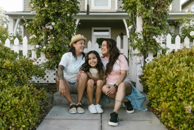 Three people, two adults and one child, sit together smiling on a front porch step, surrounded by greenery and a white picket fence.