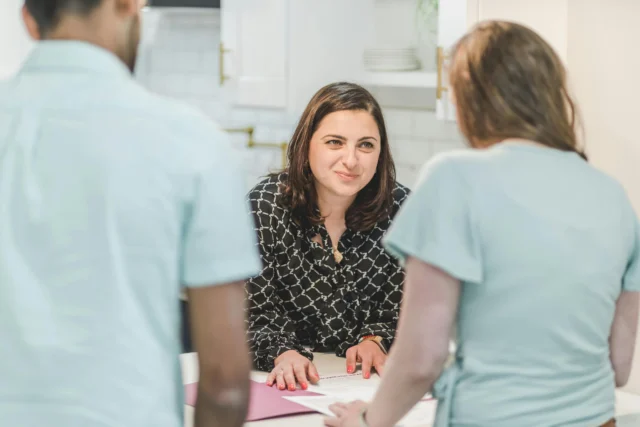 A woman stands at a kitchen counter facing two people, looking at them while resting her hands on documents spread out on the counter.