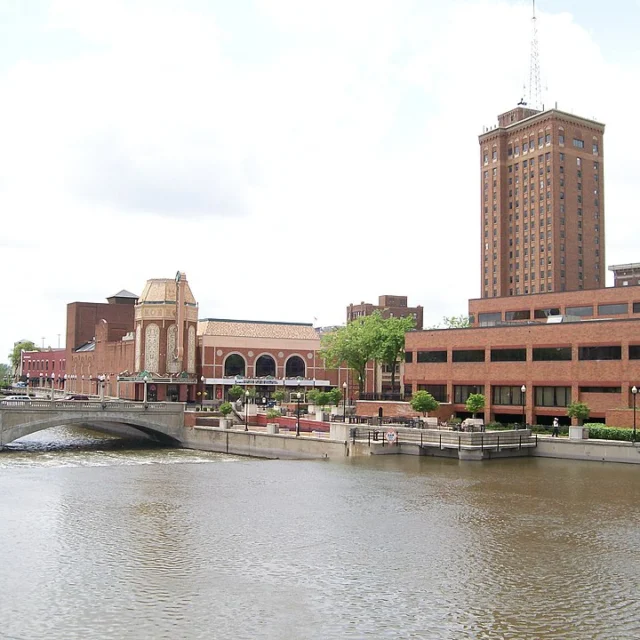 A river runs alongside brick buildings, including a tall tower, with a bridge crossing the water under a partly cloudy sky.