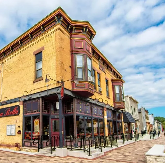 A historic yellow brick building on a street corner houses the restaurant Cucina Bella, with outdoor seating and decorative architectural details.