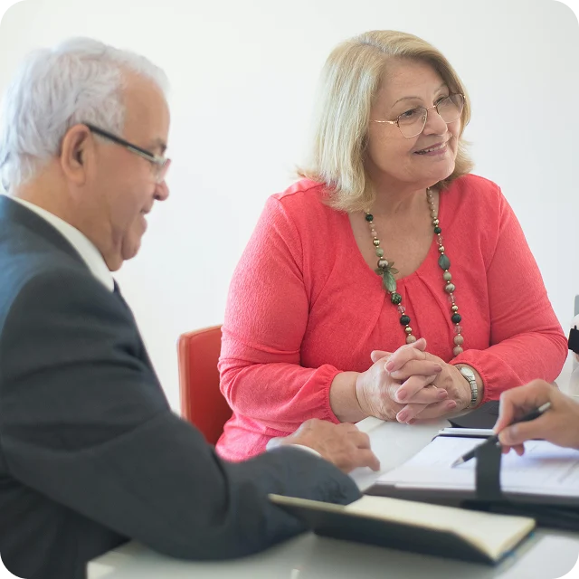 Two older adults sit at a table, listening attentively to someone off-camera who is holding a pen and clipboard, suggesting a meeting or consultation.