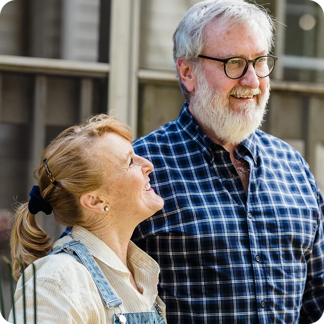 A woman smiles and looks up at a man with glasses and a beard. Both are standing outside in front of a window, dressed in casual clothes.