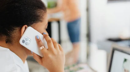 A person with blue nail polish holds a smartphone to their ear, speaking on a call while sitting at a desk with a laptop.