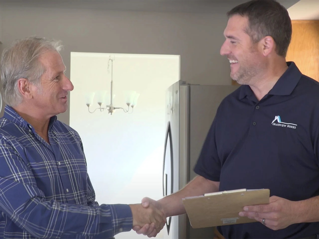 Two men shake hands indoors; one holds a clipboard and wears a collared shirt with a logo. They both appear to be smiling.