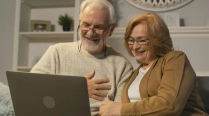 An elderly man and woman sit close together on a couch, smiling and looking at a laptop screen in a cozy, softly lit living room.