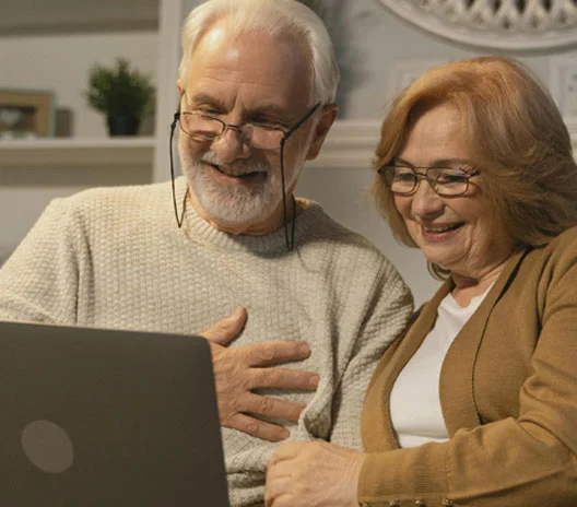 An elderly man and woman sit close together on a couch, smiling and looking at a laptop screen in a cozy, softly lit living room.