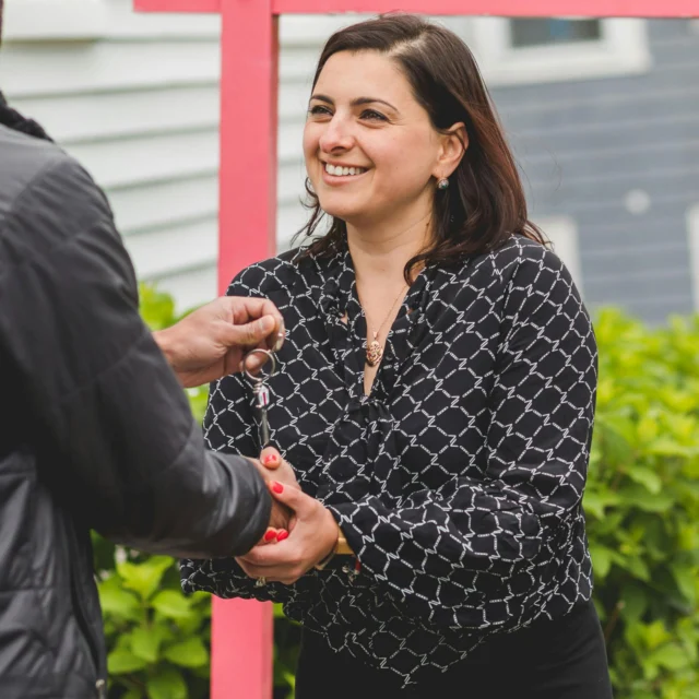 A woman smiling as she receives a set of keys from another person outdoors, with a house and greenery in the background.