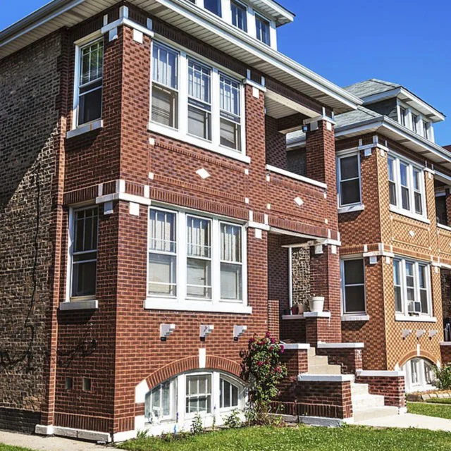 A brick two-flat residential building with white-trimmed windows, front steps, and a small garden, under a clear blue sky.