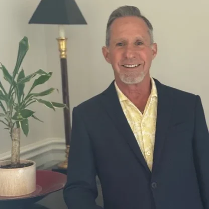 A man in a dark blazer and yellow patterned shirt stands indoors next to a potted plant and a floor lamp, smiling at the camera.