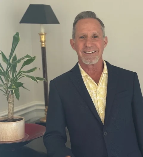 A man in a dark blazer and yellow patterned shirt stands indoors next to a potted plant and a floor lamp, smiling at the camera.