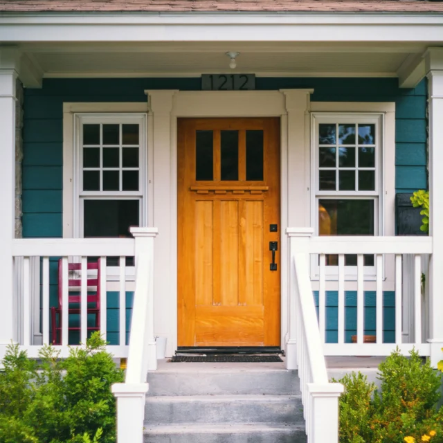 A wooden front door with a glass panel is centered between two white-trimmed windows on a teal house. The porch has white railings, with shrubs and steps leading to the entrance.