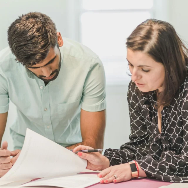 Two people review documents together at a table. The man is standing and looking down, while the woman is seated, holding a pen and focusing on the paperwork.