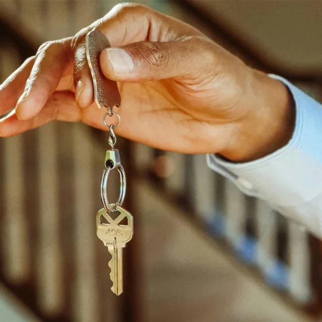 A hand holding a key on a keyring, with a staircase and banister blurred in the background.