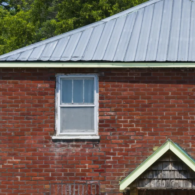 A red brick building with a single white-framed window and a metal roof, set against a background of green trees.