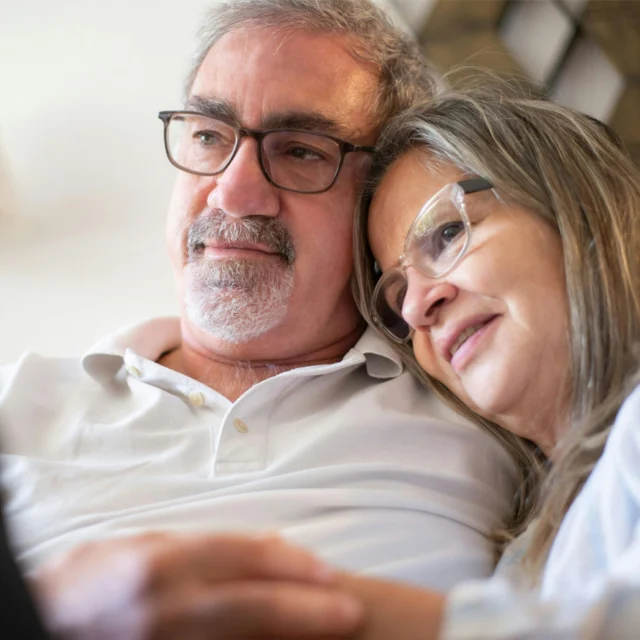 An older man and woman wearing glasses sit close together on a couch, with the woman resting her head on the man's shoulder.