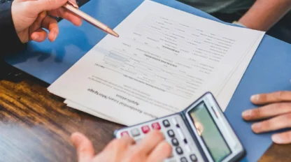 Two people sit at a desk with a laptop, reviewing documents in a blue folder; one uses a calculator while the other points at the paperwork with a pen.
