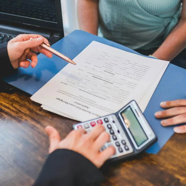 Two people sit at a desk with a laptop, reviewing documents in a blue folder; one uses a calculator while the other points at the paperwork with a pen.