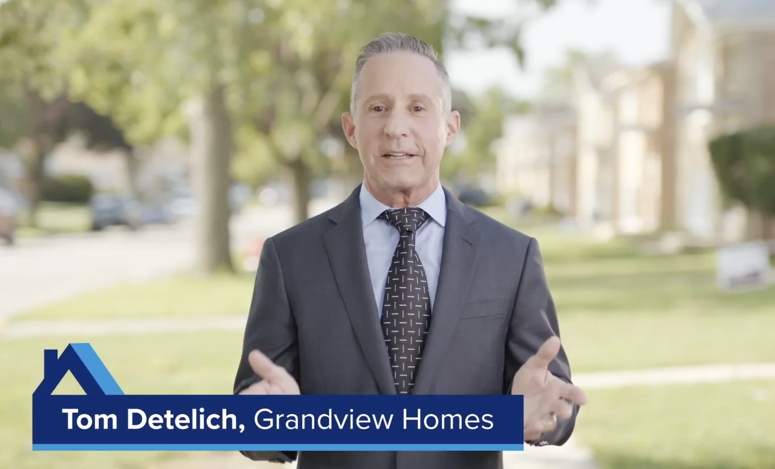 A man in a suit stands outdoors speaking, with a graphic text overlay that reads "Tom Detelich, Grandview Homes." Trees and houses are visible in the blurred background.
