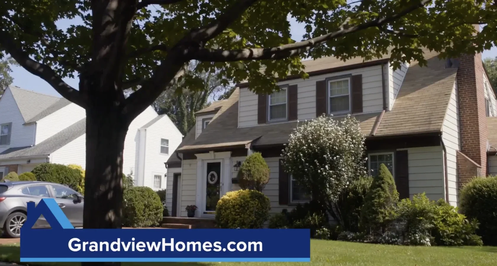 A two-story suburban house with brown shutters, surrounded by trees and shrubs, with "GrandviewHomes.com" displayed on a blue banner in the foreground.