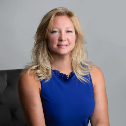 A woman with blonde hair in a blue sleeveless dress sits on a dark chair, posing with hands resting on her knee against a plain gray background.