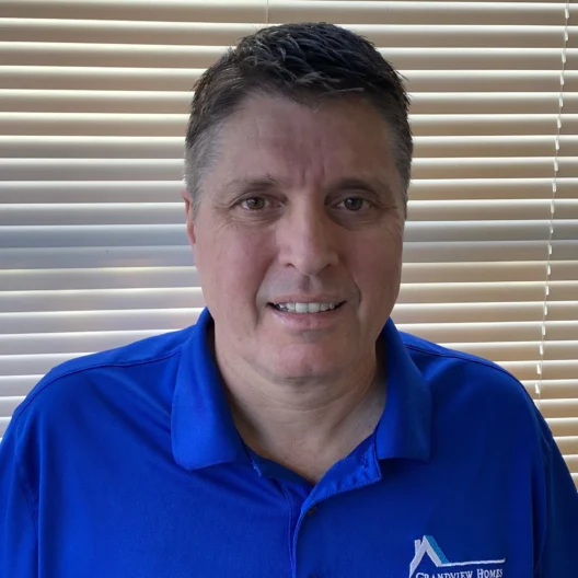 A man in a blue "Grandview Homes" polo shirt stands in front of window blinds, looking at the camera and smiling slightly.