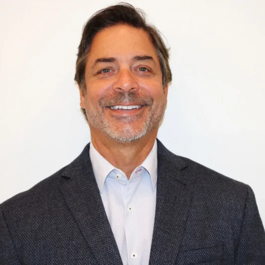 A middle-aged man with short brown hair and a beard wearing a dark textured blazer and light shirt, smiling in front of a plain white background.