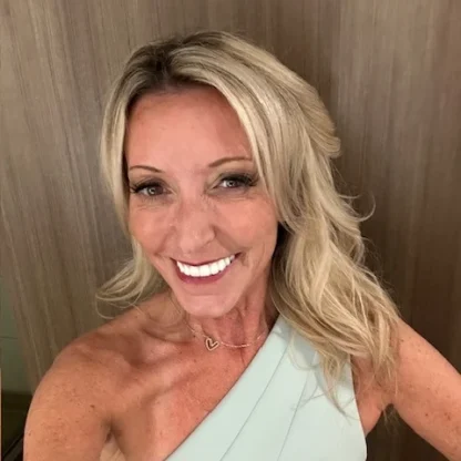 A woman with long blonde hair, wearing a one-shoulder light-colored top, smiles at the camera in front of a wooden background.