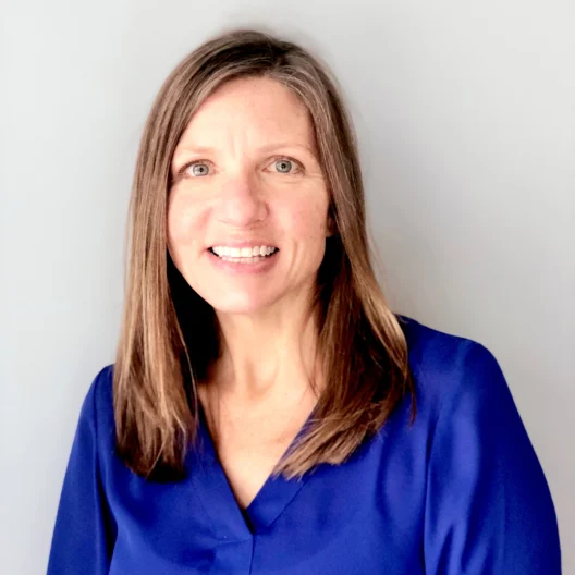 A woman with straight brown hair wearing a blue blouse is smiling at the camera against a plain light grey background.