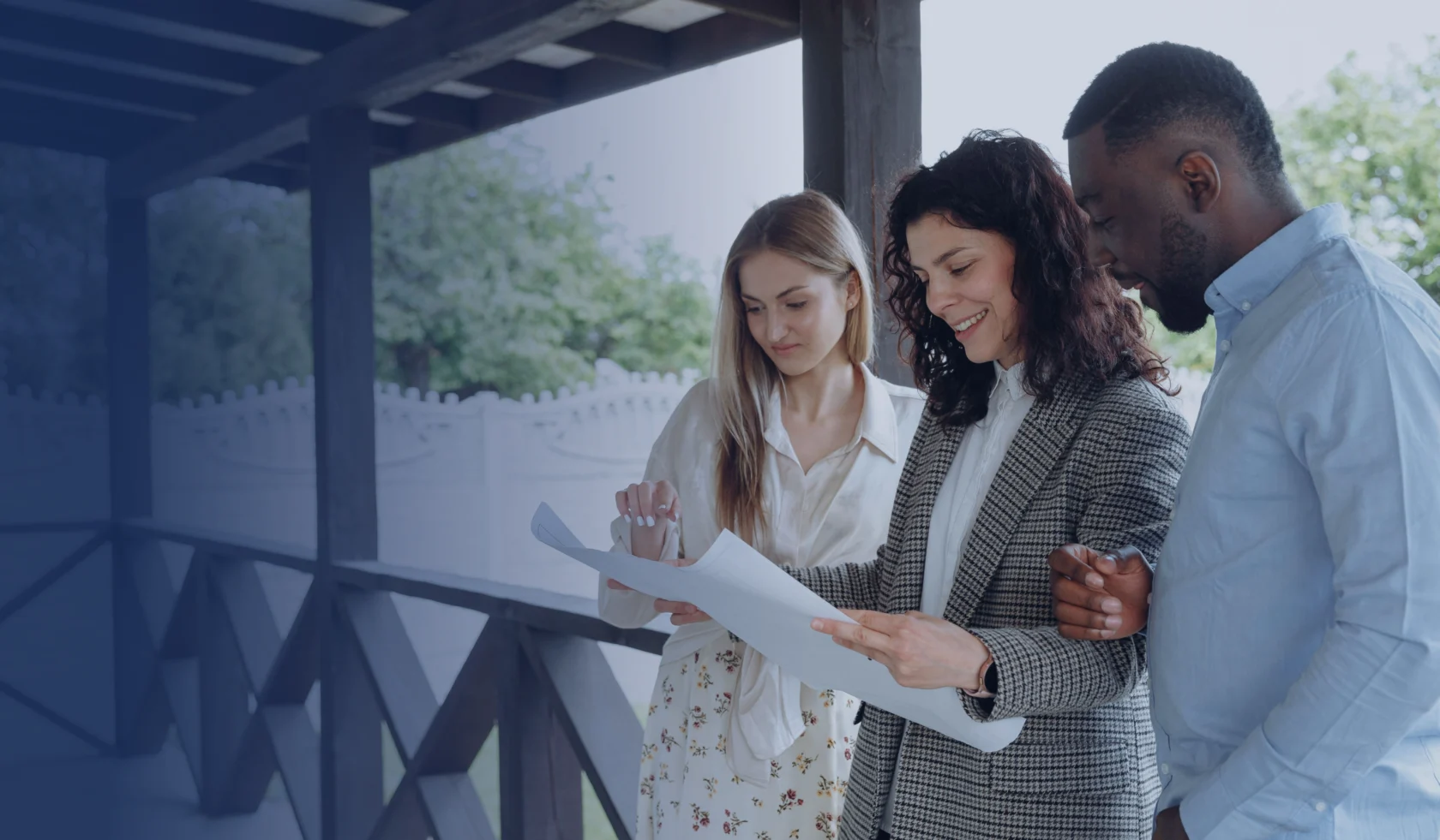Three people stand together on a porch, looking at documents and discussing something, with a wooden fence and greenery in the background.