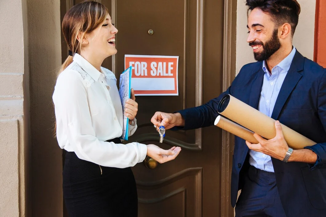 A man hands a woman a set of keys in front of a door with a "For Sale" sign. Both are dressed in business attire and appear to be engaged in a real estate transaction.