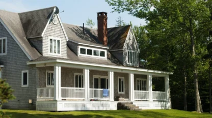 Two-story gray house with a large front porch, white railings, and dormer windows, surrounded by green grass and trees on a sunny day.