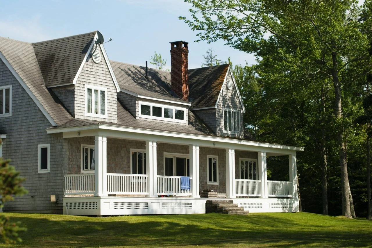 Two-story gray house with a large front porch, white railings, and dormer windows, surrounded by green grass and trees on a sunny day.