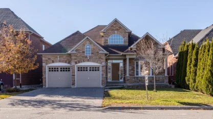Two-story suburban house with brick exterior, double garage doors, a covered porch, and a small front yard with grass and trees.