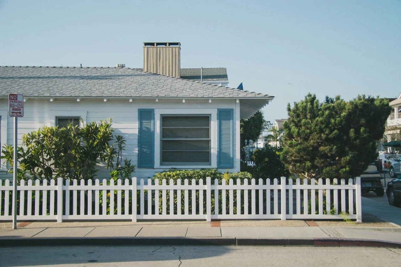 Single-story house with blue shutters, white picket fence, green bushes, and a "No Parking" sign along the sidewalk under a clear sky.