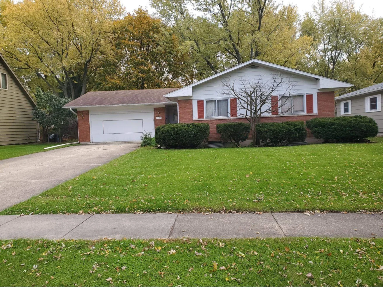 Single-story brick and white house with an attached garage, trimmed bushes, a young tree in front yard, and green lawn bordered by sidewalk.