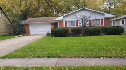 Single-story brick and white house with an attached garage, trimmed bushes, a young tree in front yard, and green lawn bordered by sidewalk.