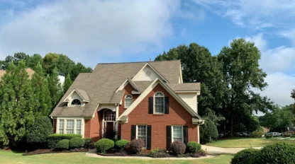 A two-story suburban house with red brick and beige siding, surrounded by neatly trimmed bushes, trees, and a well-kept lawn under a partly cloudy sky.