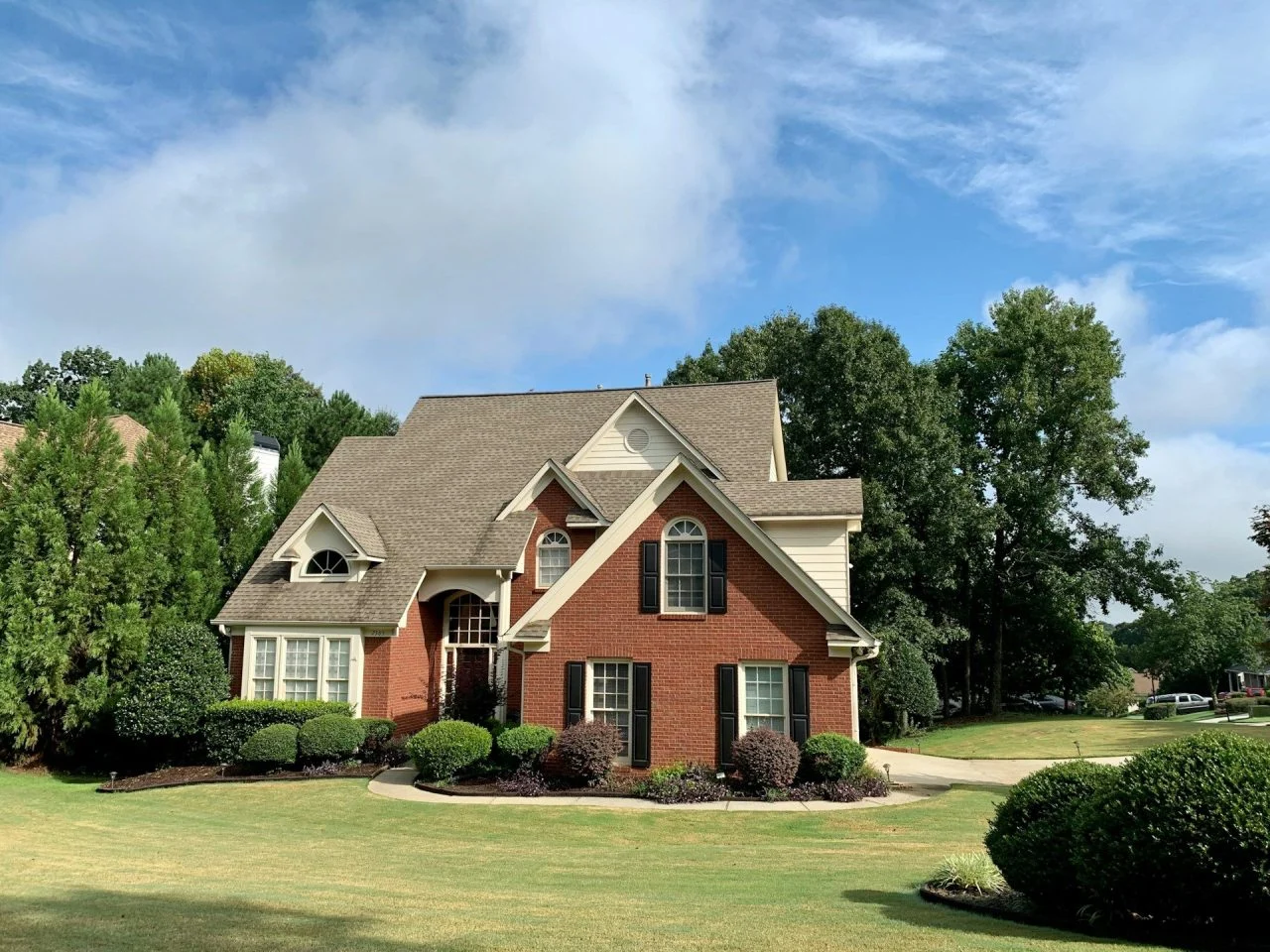 A two-story suburban house with red brick and beige siding, surrounded by neatly trimmed bushes, trees, and a well-kept lawn under a partly cloudy sky.