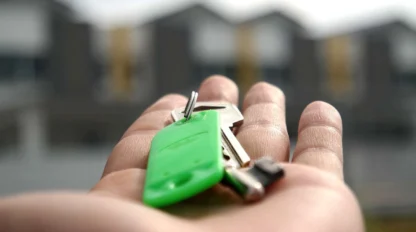 A close-up of a hand holding a set of house keys with a green key tag, with blurred houses in the background.