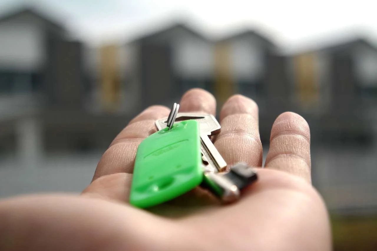 A close-up of a hand holding a set of house keys with a green key tag, with blurred houses in the background.