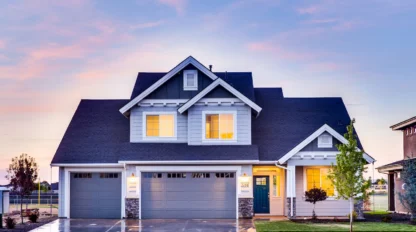 A two-story suburban house with three garage doors, lit windows, and a wet driveway at sunset.