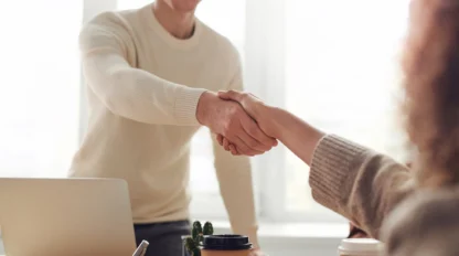 Two people are shaking hands across a desk with a laptop, notepad, and coffee cups, suggesting a business meeting or agreement in a well-lit office setting.