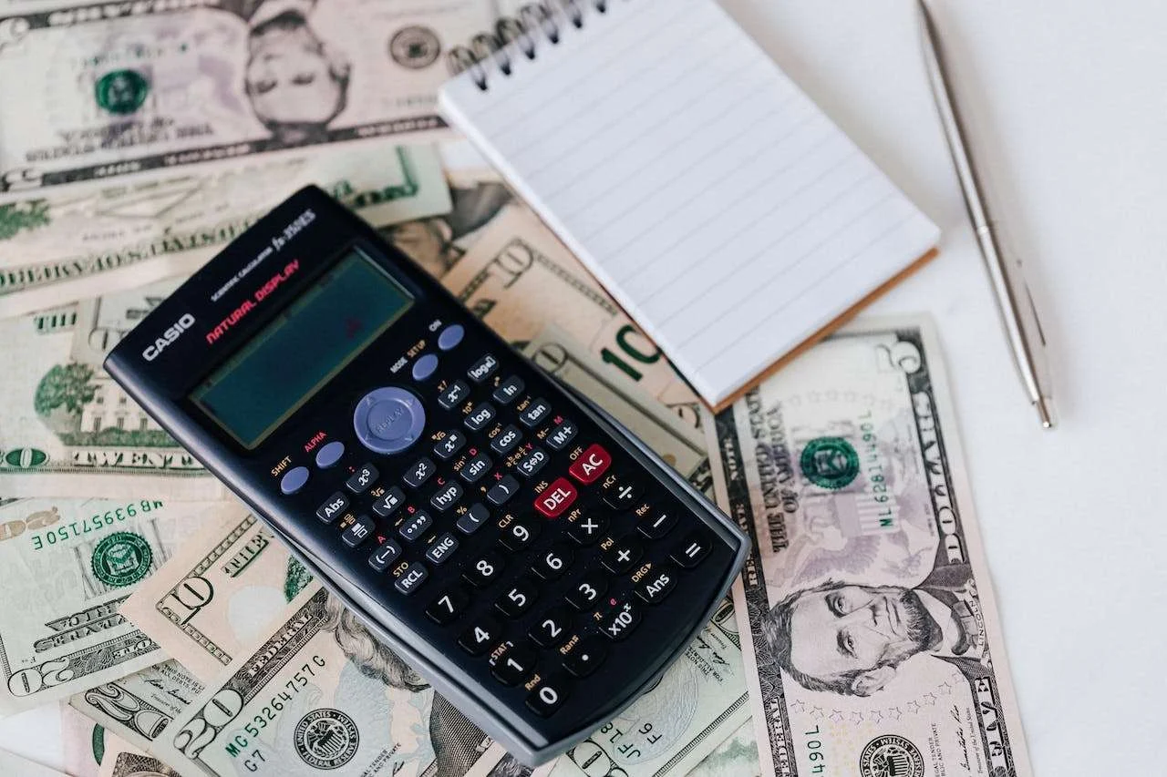 A scientific calculator, a notepad, and a pen are placed on top of scattered U.S. dollar bills on a white surface.