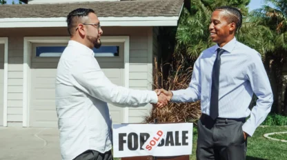 Two men shake hands in front of a house with a "For Sale" sign marked "Sold," indicating a completed real estate transaction.