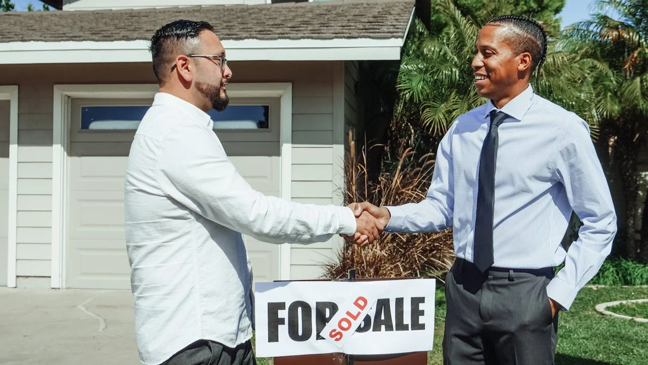 Two men shake hands in front of a house with a "For Sale" sign marked "Sold," indicating a completed real estate transaction.