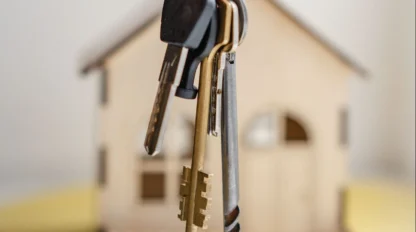 A hand holds a set of house keys in front of a small model house with a blurred background.