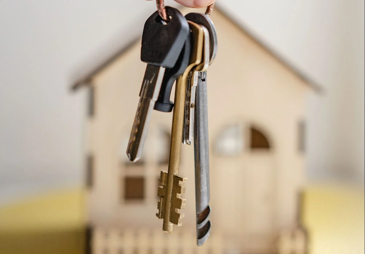 A hand holds a set of house keys in front of a small model house with a blurred background.