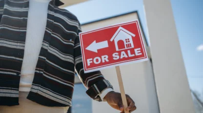 A person holding a red "For Sale" sign with a house icon and arrow, standing in front of a modern building.