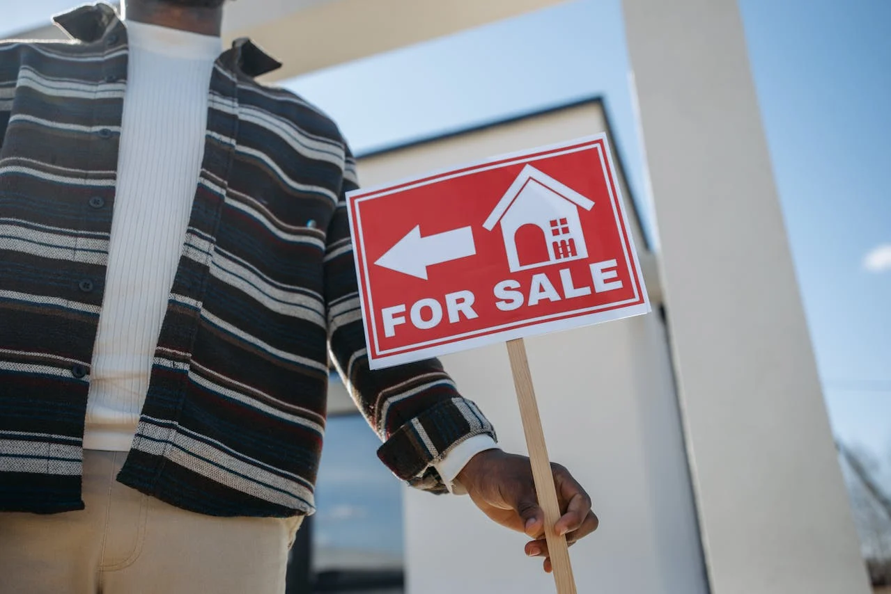 A person holding a red "For Sale" sign with a house icon and arrow, standing in front of a modern building.