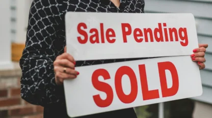 Person holding "Sale Pending" and "SOLD" signs in front of a house, indicating a property has been sold.